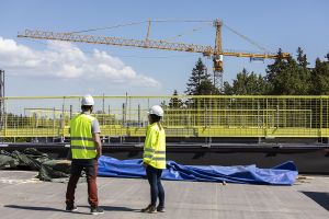 Två SKB-medarbetare står på taket till ett hus under byggnation. I bakgrunden en lyftkran som reser sig över en skogsdunge.