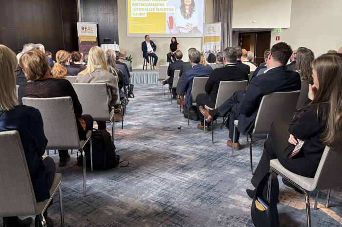 Stor lokal där man ser ryggarna på många åhörare och i mitten en scen på långt avstånd, där två personer samtalar.