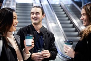 Tre unga personer i glatt samtal. De dricker kaffe vid rulltrappor.